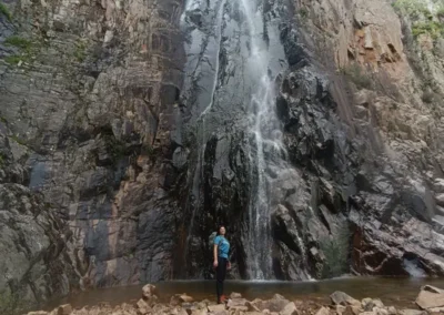 Veronica Murgia in piedi su un terreno roccioso davanti a una cascata che scende lungo una parete di roccia.