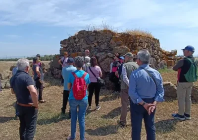 Gruppo di escursionisti riuniti attorno a Veronica Murgia di Vemu Inclusive Hikes durante una visita a un nuraghe in pietra, immerso in un paesaggio rurale.