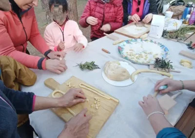 Gruppo di persone, tra cui donne e bambini, impegnate in un laboratorio di pasta fresca all'aperto, in un bosco.