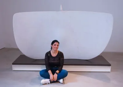 Donna seduta a terra sorridente davanti a una grande scultura in marmo bianco dalla forma astratta, esposta in un ambiente museale.