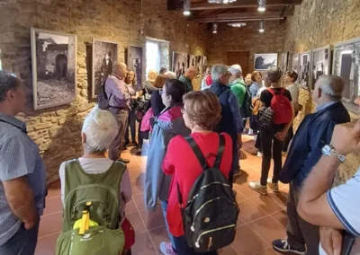 Gruppo di persone durante una visita guidata in una sala con pareti in pietra, osservando fotografie storiche esposte.